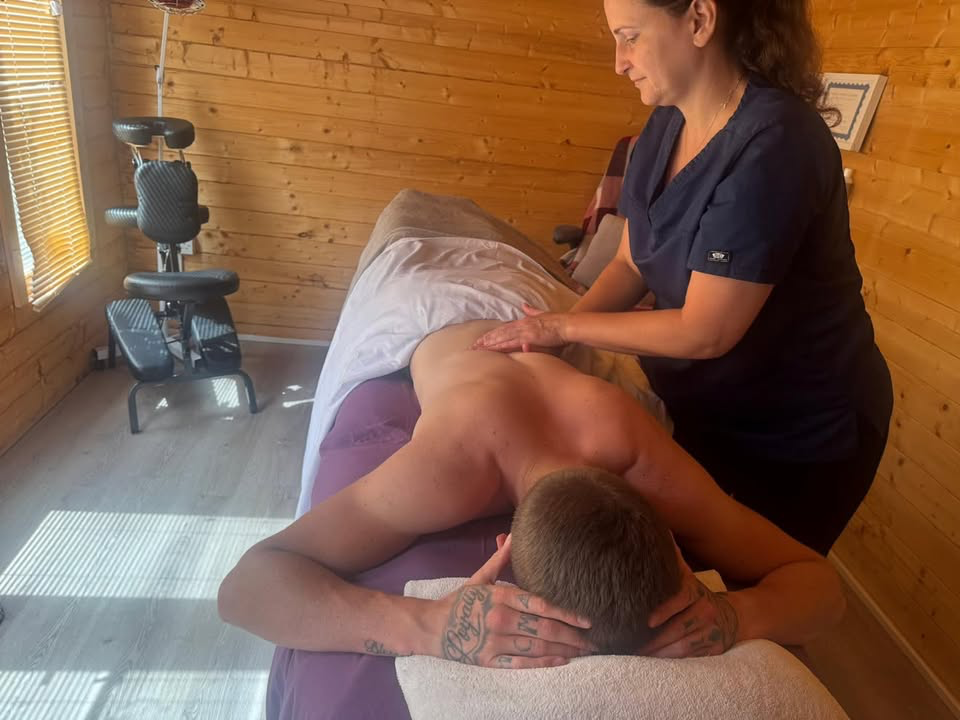 Diane treating a client on the massage couch inside the wood-lined garden cabin, with the portable massage chair visible in the background